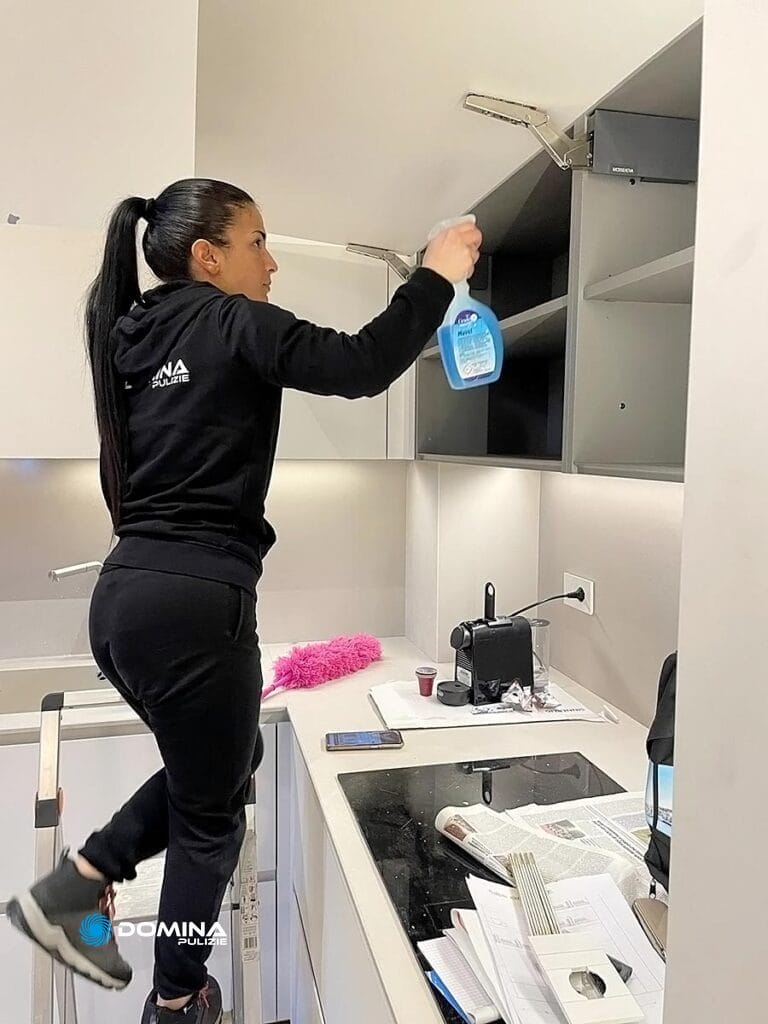 Una persona di Domina Pulizie sta pulendo diligentemente l'interno di un mobile da cucina mentre si trova su una piccola scala, circondata dai prodotti per la pulizia Domina.