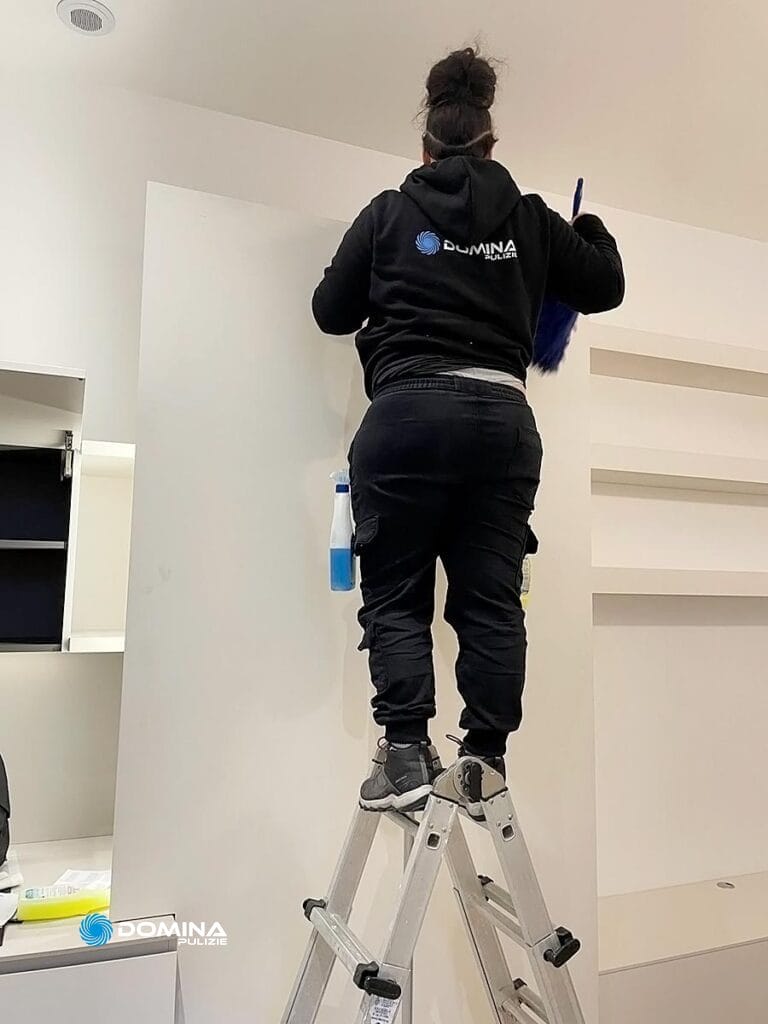 Una persona con una felpa nera con il logo "Domina" sul retro è in piedi su una scala a pioli mentre pulisce o spolvera uno scaffale bianco, vista da dietro.