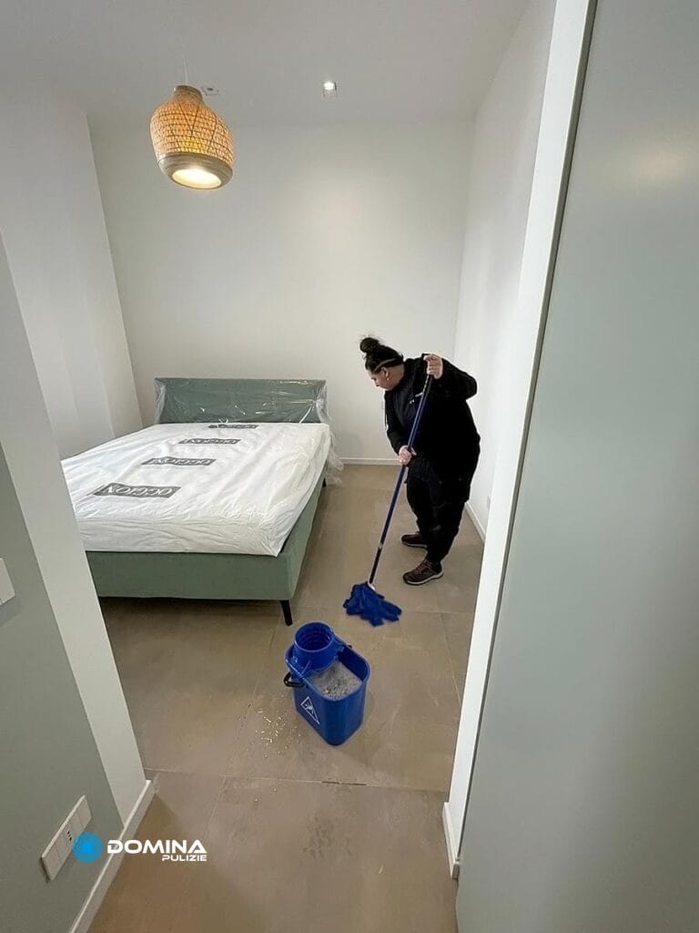 Una persona che pulisce il pavimento di una camera da letto ordinata e arredata in modo minimale con un letto appena rifatto, esemplificando gli standard impeccabili di Domina Pulizie Appartamenti Seregno.