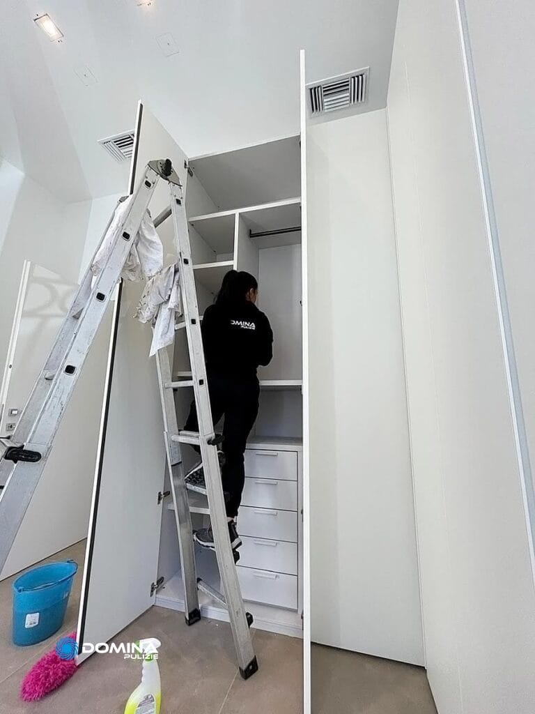 Una persona con divisa di Domina Pulizie o pulendo uno scaffale alto in una moderna cabina armadio bianca, utilizzando una scala a pioli in alluminio, con Pulizie Appartamenti