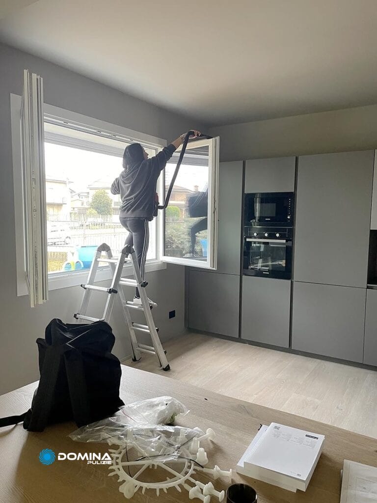 Una persona su una scala pulisce una finestra in una cucina moderna con mobili grigi ed elettrodomestici in acciaio inossidabile per Impresa di Pulizie Monza.