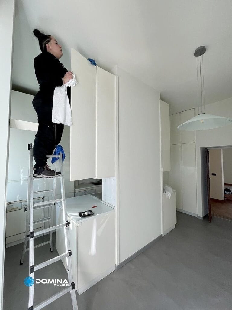 Donna su una scala che pulisce con attenzione la parte alta di un alto mobile da cucina per l'Impresa di Pulizie Domina Pulizie Monza.