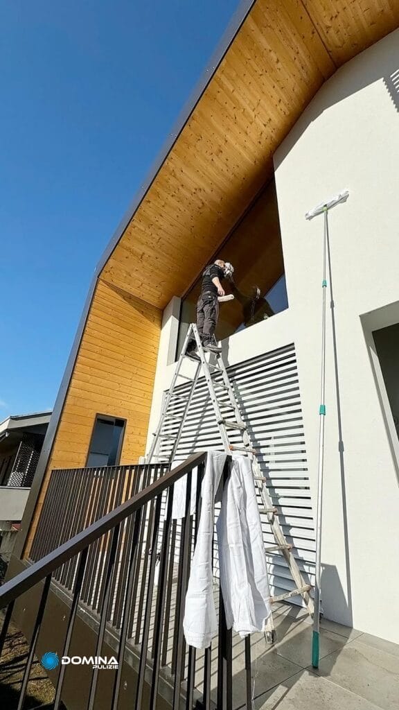 Una persona in tenuta da lavoro in piedi su una scala mentre pulisce delle vetrate all'esterno di un edificio moderno con tetto curvo in legno per Impresa di Pulizie a Monza.