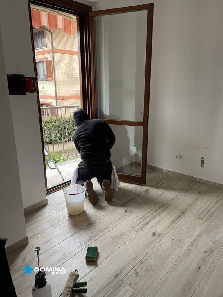 Persona inginocchiata mentre pulisce o installa una porta di vetro in una casa ad Arluno, con attrezzi e materiali per la pulizia nelle vicinanze.