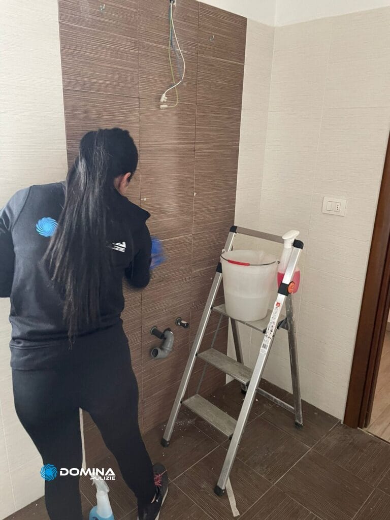 Una persona effettua le pulizie post ristrutturazione di una casa ad Arluno, pulendo una parete piastrellata accanto a una scala con prodotti
