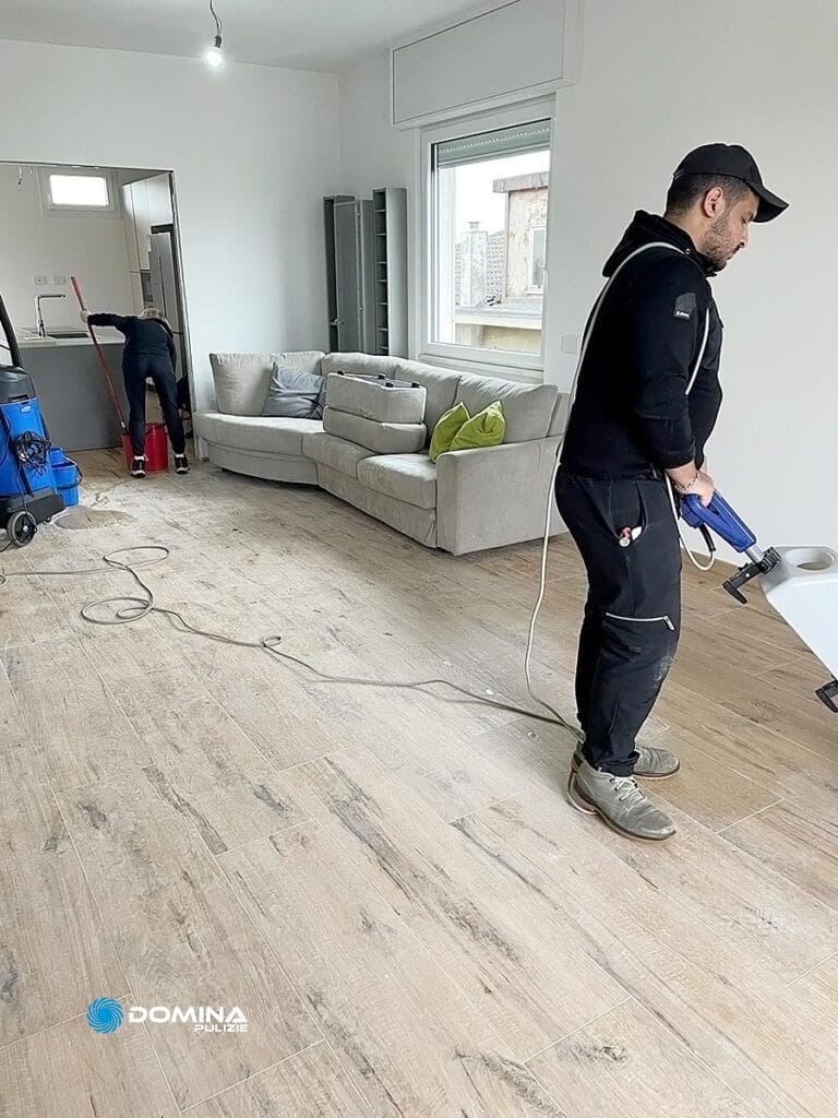 Un uomo di Domina Pulizie sta pulendo il pavimento del soggiorno con un'aspirapolvere professionale mentre un'altra persona lavora in background.