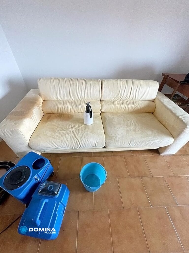 Un divano color crema con sopra una bottiglia, affiancato da un aspirapolvere blu e un secchio su un pavimento piastrellato, che suggerisce una sessione di pulizia di Domina Pulizie.