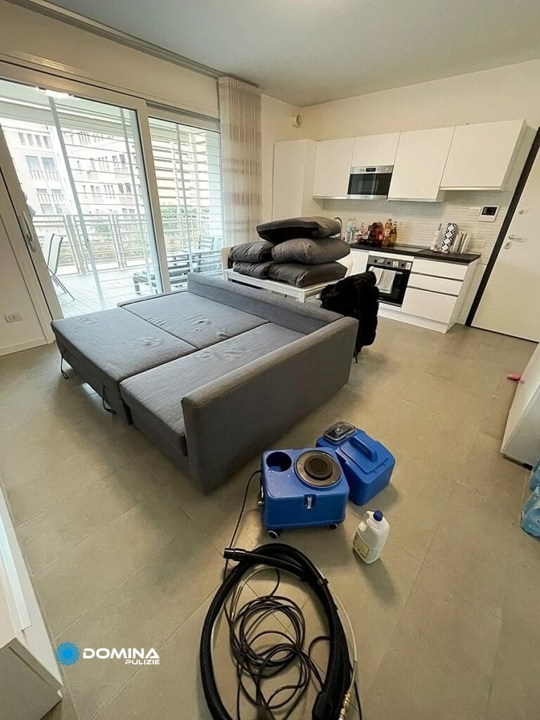 Soggiorno di un appartamento moderno con cucina a vista, divano componibile grigio in fase di "Lavaggio divani Bollate" e vari oggetti per la casa sparsi sul pavimento.