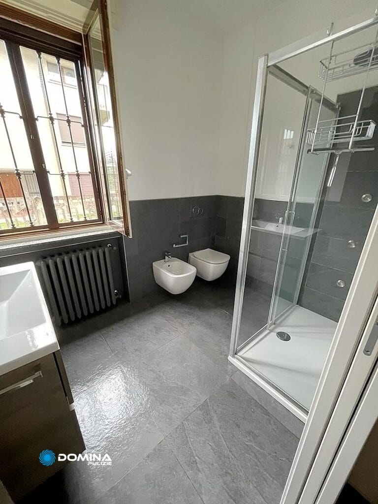 Bagno moderno con cabina doccia, WC sospeso, bidet e lavandino vicino ad una finestra con vista sull'esterno, curato da Domina Pulizie.