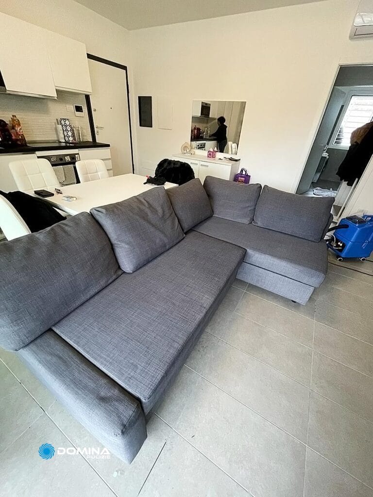 Soggiorno moderno con divano grigio a L, lavato da Domina Pulizie, e sullo sfondo un ordinato angolo cottura bianco.