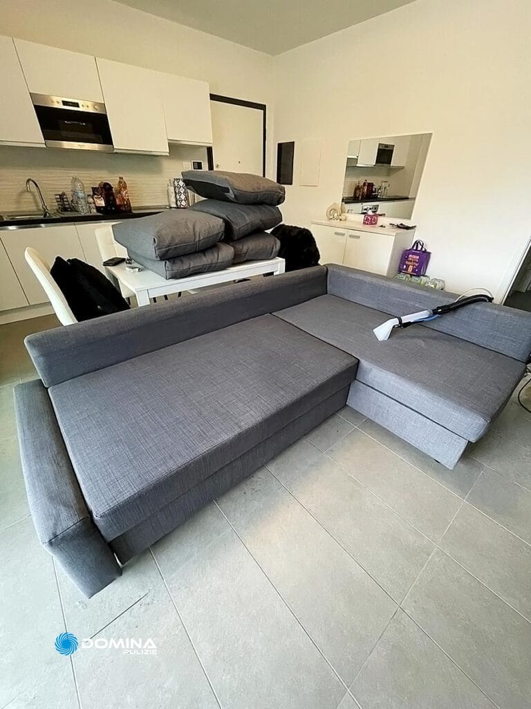 Un soggiorno ordinato con un divano componibile grigio, che beneficia del servizio di lavaggio divani di Domina Pulizie, e una moderna zona cucina sullo sfondo.