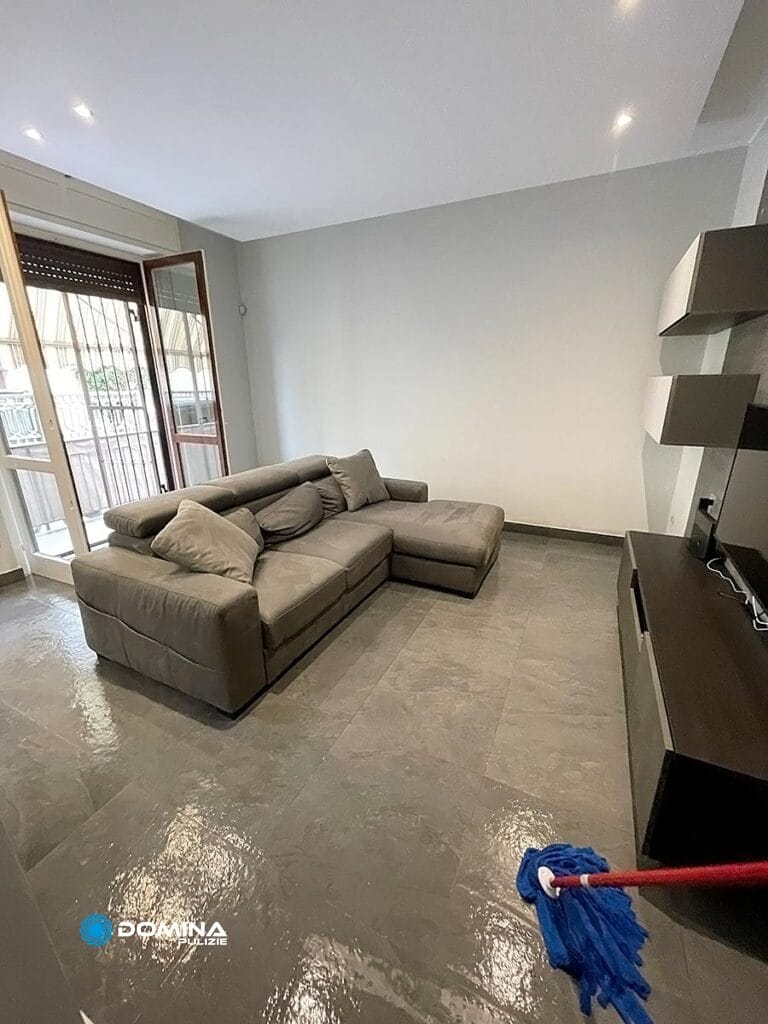 Un soggiorno pulito e moderno con un divano componibile grigio e un pavimento piastrellato lucido, appena lavato da Domina Pulizie.