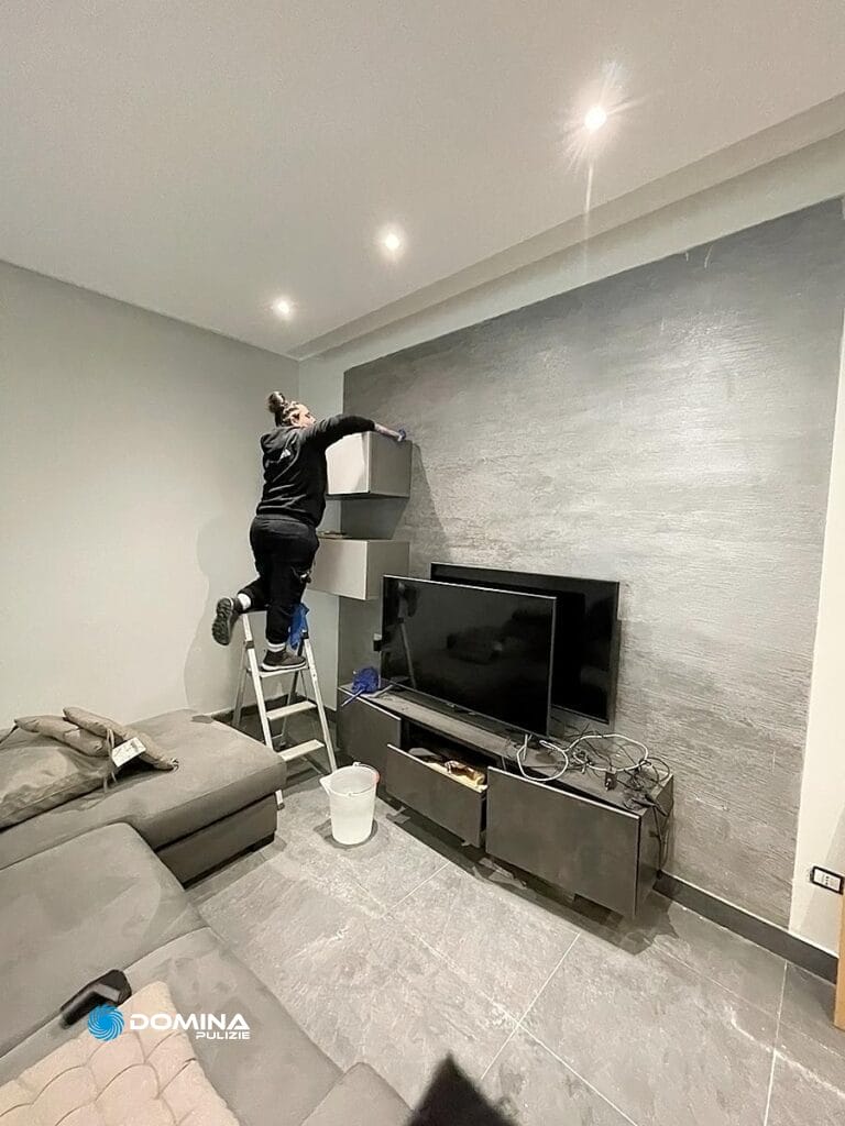 Persona su una scala di Domina Pulizie che pulisce uno scaffale sopra un televisore in un soggiorno moderno.