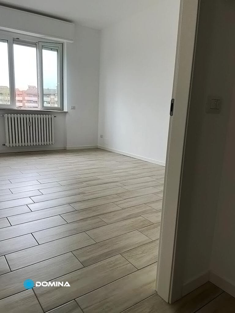 Una stanza vuota con pareti bianche, una grande finestra, pavimento in legno chiaro e mantenuta meticolosamente da Domina Pulizie.