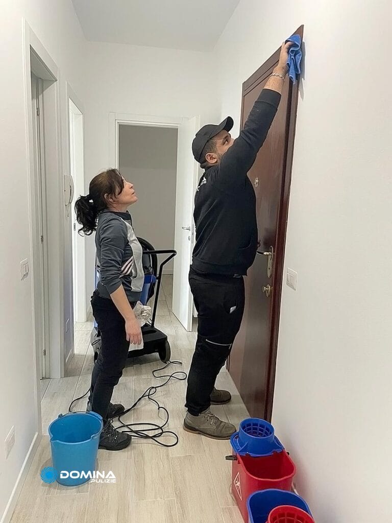 Due persone si occupano della pulizia post cantiere di una porta a Milano, una asciugando il bordo superiore mentre l'altra osserva.