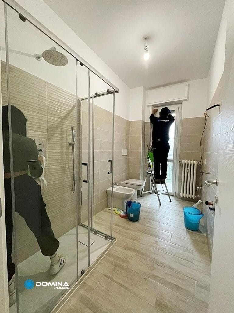 Una persona su una scala che esegue la manutenzione e pulizia post cantiere Milano lavora in un bagno ben illuminato con cabina doccia in vetro.