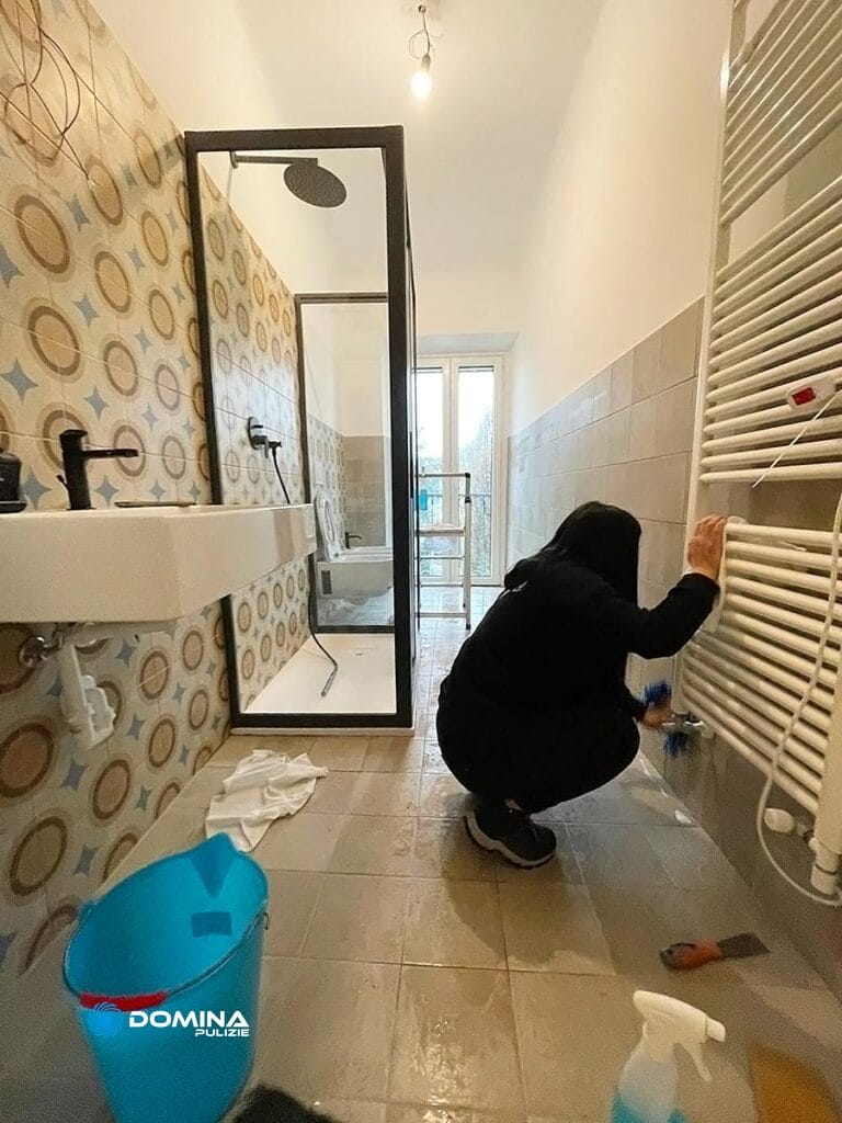 Persona vestita di nero che pulisce il pavimento del bagno vicino a un termosifone, con prodotti per la pulizia e un secchio blu nelle vicinanze, nell'ambito del servizio di pulizia post-costruzione di Domina Pulizie a Bollate.