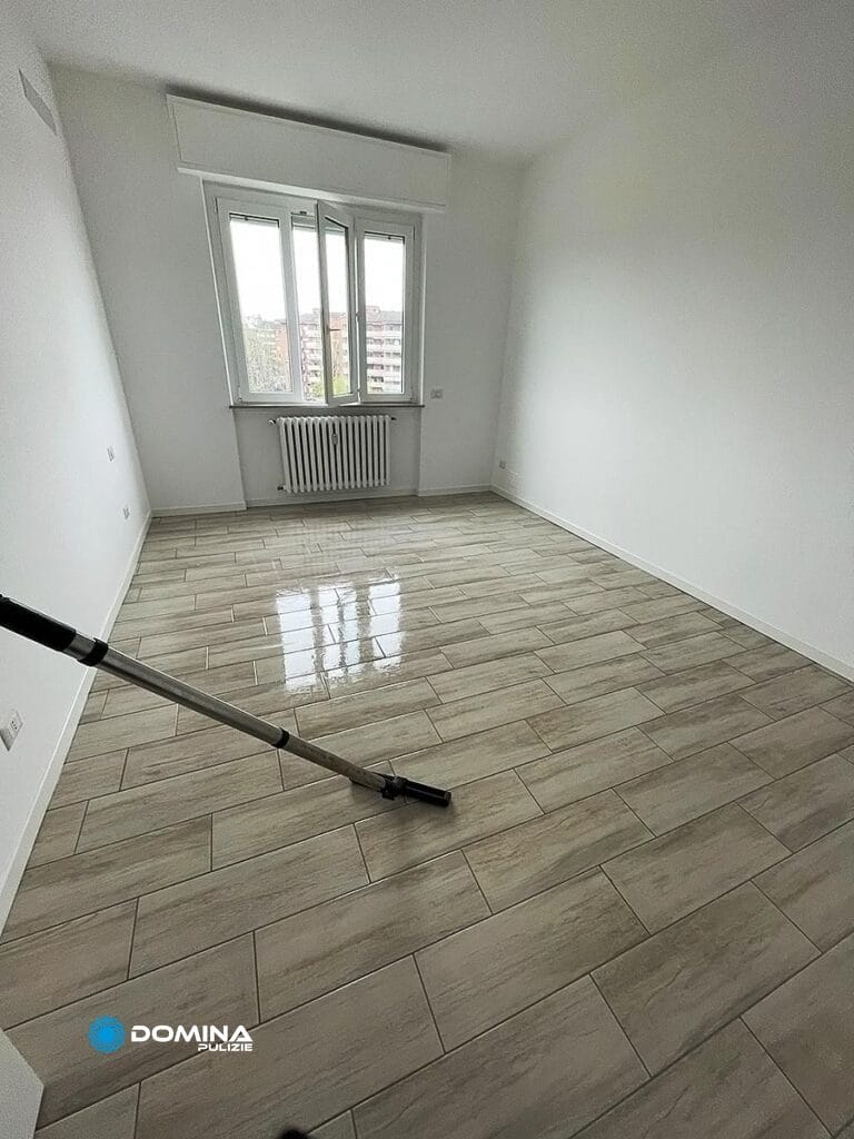 Stanza vuota con pavimento piastrellato pulito con uno spazzolone Domina Pulizie vicino a una finestra da cui entra luce naturale.