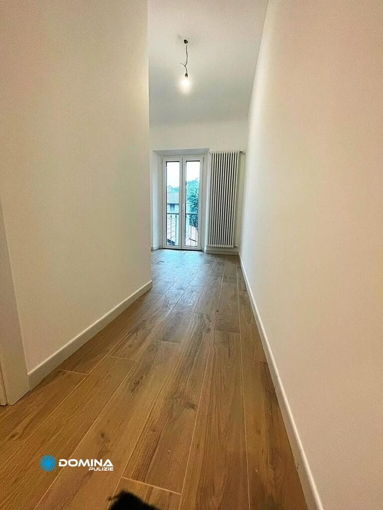 Un corridoio stretto e vuoto, con pavimento in legno, conduce ad una stanza con porta-balcone, illuminata da luce naturale e mantenuta meticolosamente da Domina Pulizie.