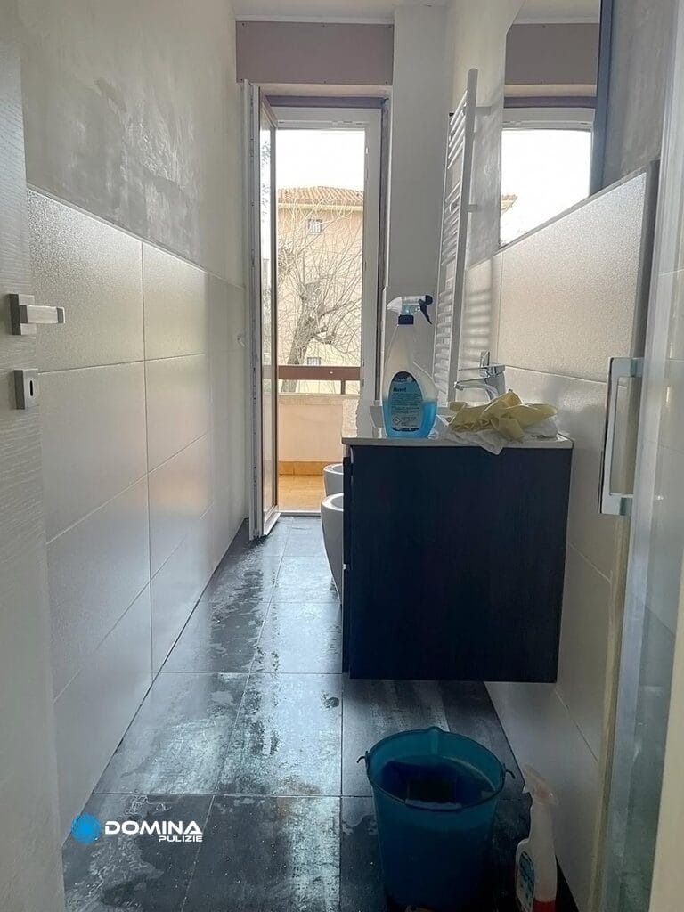 Interno del bagno con luce naturale, caratterizzato da un secchio blu su un pavimento bagnato e prodotti per la pulizia Domina Pulizie sul ripiano del lavandino.
