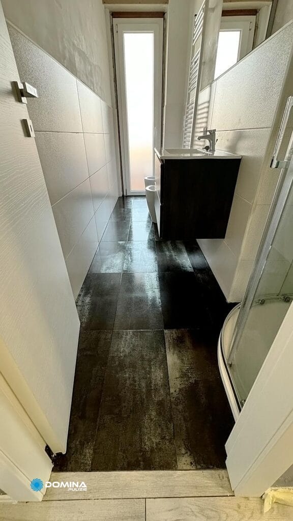 Un bagno stretto con la doccia a destra, un lavabo nero davanti e un pavimento piastrellato che conduce ad una finestra con le persiane leggermente socchiuse, curata da Domina Pulizie.