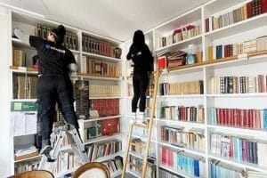 Due persone di Domina Pulizie puliscono gli scaffali superiori di una libreria bianca dal pavimento al soffitto in una stanza ben illuminata vicino a Piazzale Loreto Milano