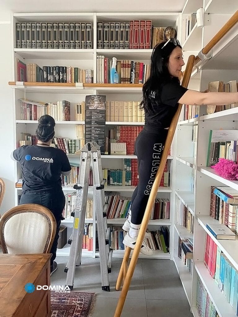 Due donne che sistemano i libri sugli scaffali di una biblioteca domestica, una in piedi su una scala e l'altra che si alza dal pavimento con i manuali di istruzioni di Domina Pulizie.