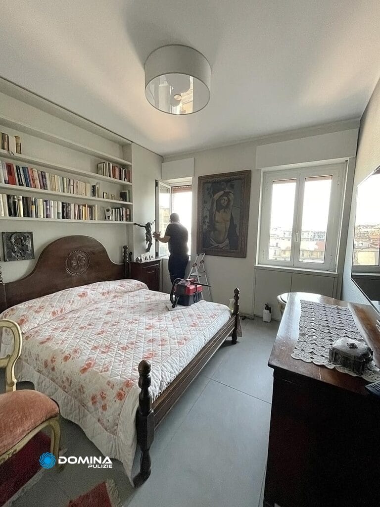 Una donna è in piedi accanto a una finestra in una camera da letto ben illuminata, piena di un grande letto, librerie, opere d'arte e mobili vintage, meticolosamente mantenuti da Domina Pulizie.