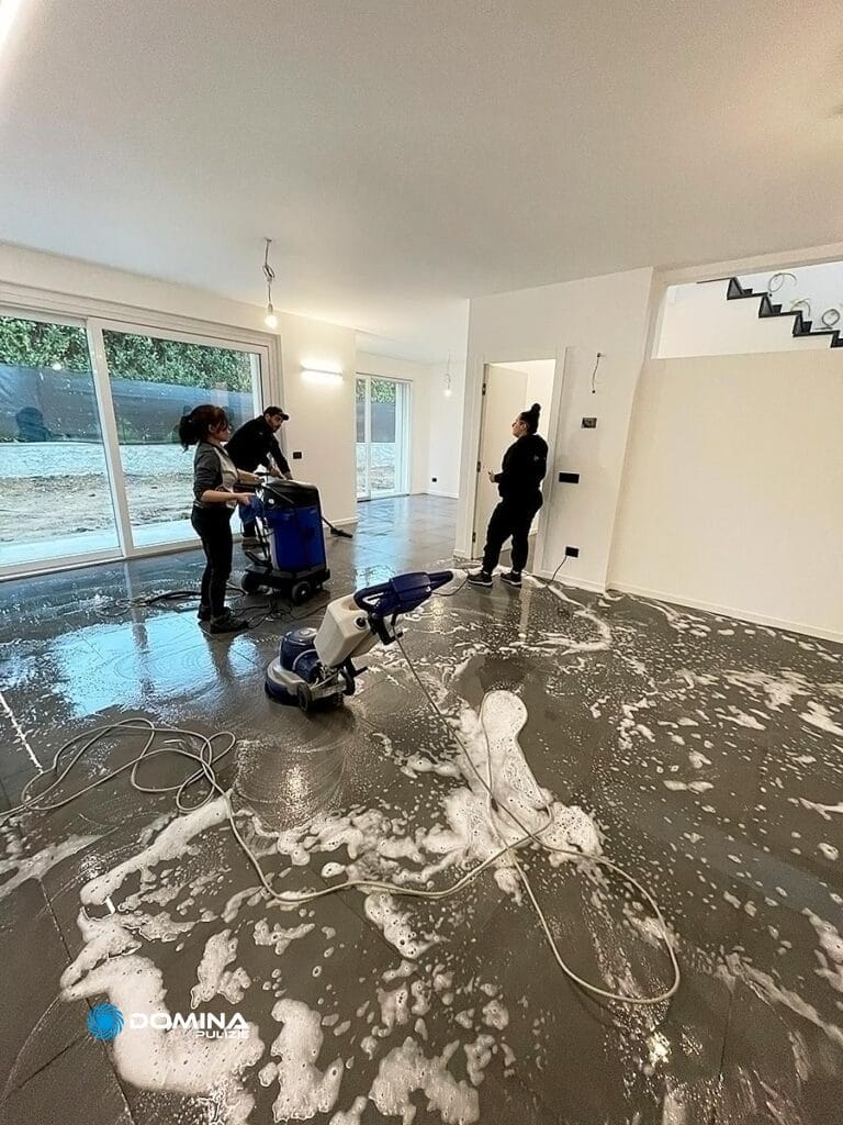 Tre persone dell'impresa di pulizie Domina puliscono una stanza bianca con grandi finestre, utilizzando una lavasciuga pavimenti e panni, con acqua saponata sul pavimento.