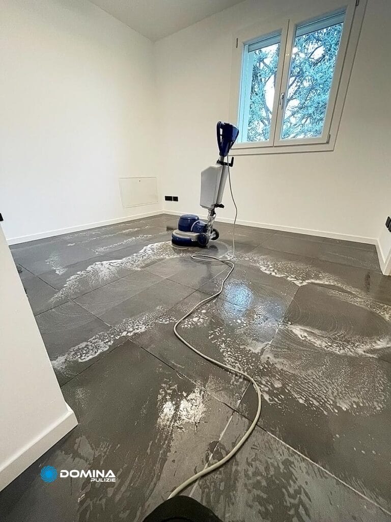 Una macchina lavasciuga pavimenti pulisce una stanza piastrellata grigia, con acqua saponata sul pavimento e una vista innevata attraverso la finestra, nell'ambito di una "Pulizia fine cantiere Cador"