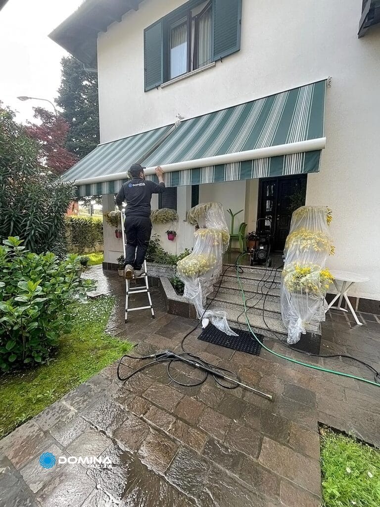 Una persona su una scala esegue la pulizia tende da esterna, con le piante circostanti coperte da sacchetti di plastica per protezione.