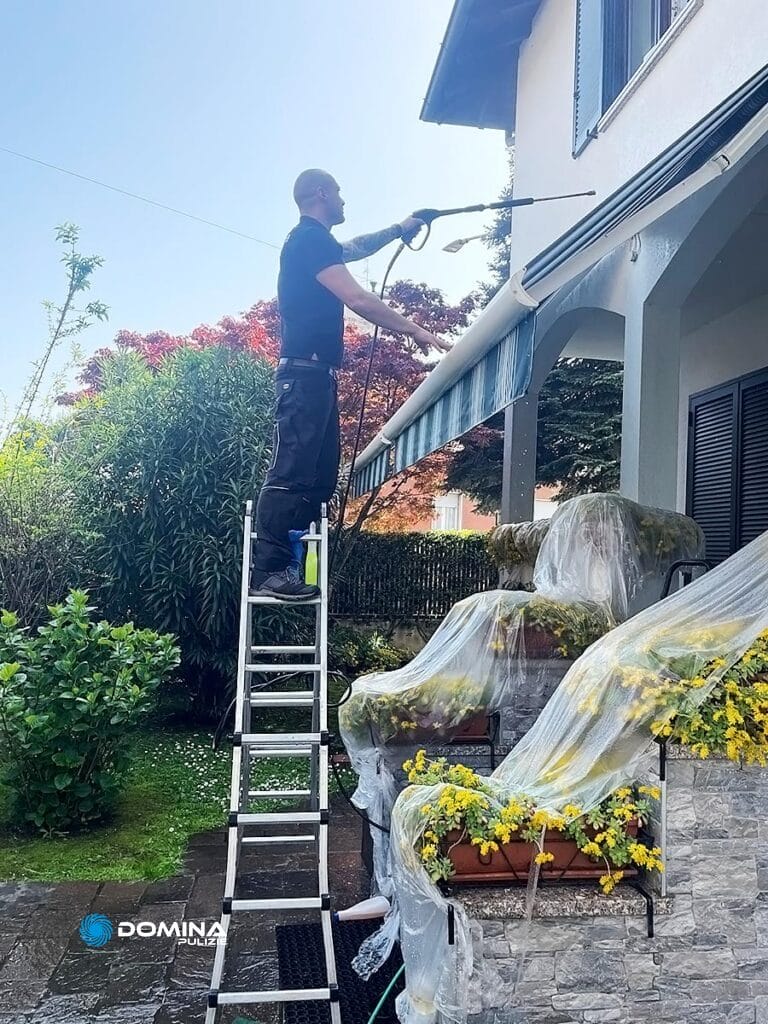 Un uomo su una scala pulisce le tende da esterno di una casa con un getto d'acqua ad alta pressione fornito dall'Impresa di pulizie Domina, con le piante coperte per protezione.