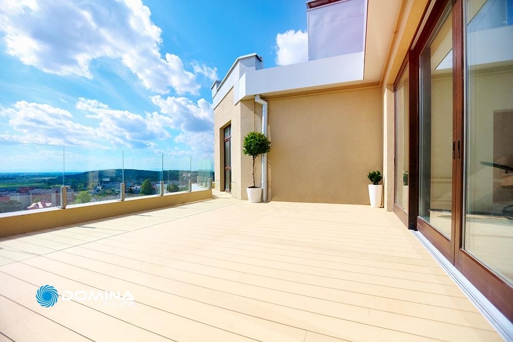 Spazioso patio sul tetto con pavimento in legno, porte in vetro e vista panoramica su una città sotto un cielo azzurro e soleggiato, mantenuto con pulizia professionale.