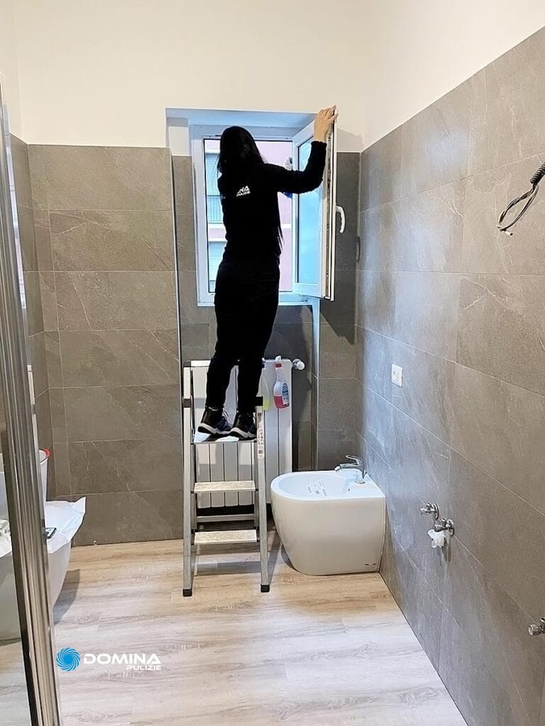 Una persona vestita di nero è in piedi su una scala mentre si allunga per aprire una finestra in un bagno con pareti piastrellate grigie e pavimento in legno chiaro, probabilmente eseguendo pulizie post ristrutturazione.