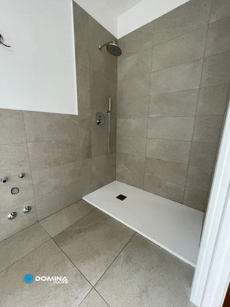 Una moderna cabina doccia piastrellata con soffione a pioggia e attacco per doccia a mano, in contrasto con piastrelle beige, con piatto doccia bianco, perfetta per completare le pulizie casa nuova.