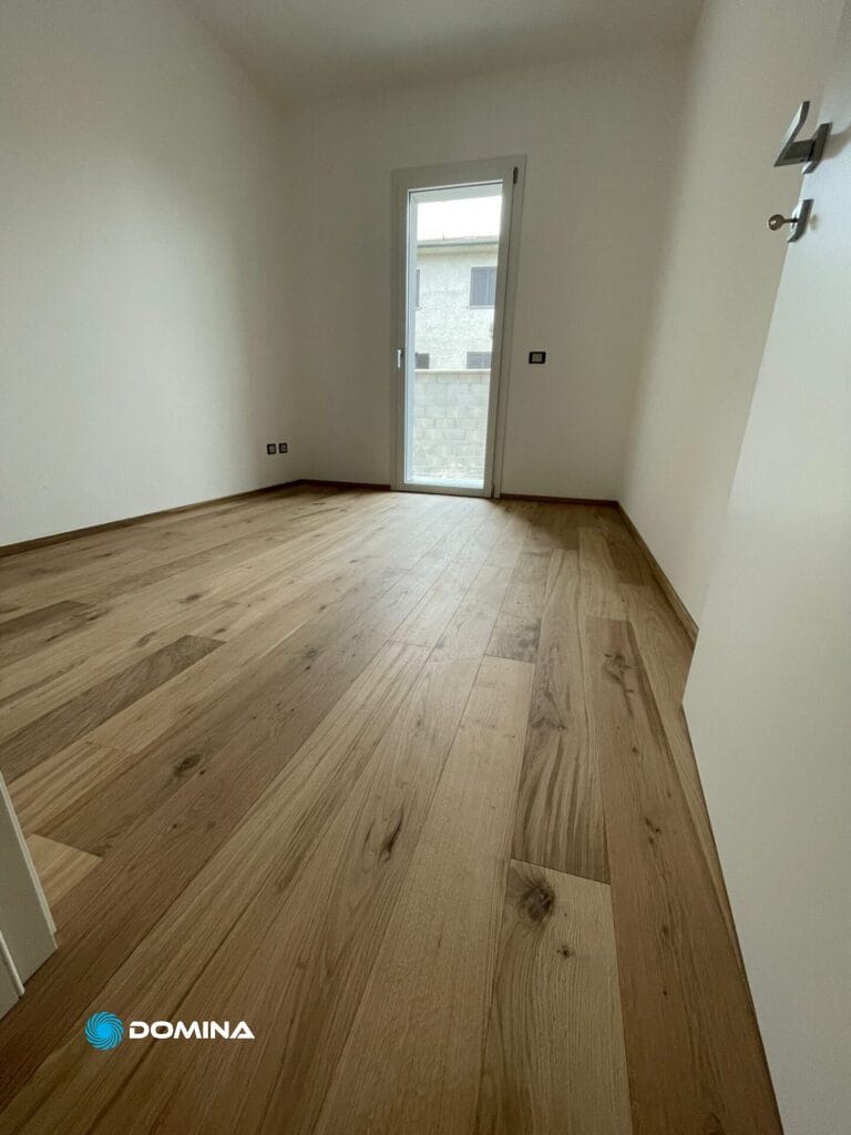 Una stanza piccola e vuota con pareti bianche e pavimento in legno, perfetta per le pulizie casa nuova. Una porta chiusa a sinistra ed una porta finestra in fondo alla stanza permettono la luce naturale proveniente dall'esterno, rendendola ideale per i servizi Domina Pulizie.