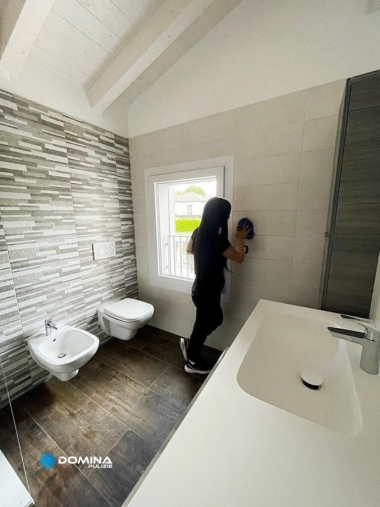 Persona che pulisce i rivestimenti in un bagno moderno con una grande finestra, lavandino, wc e bidet: pulizie approfondite nel cantiere Malnate di Domina Pulizie.