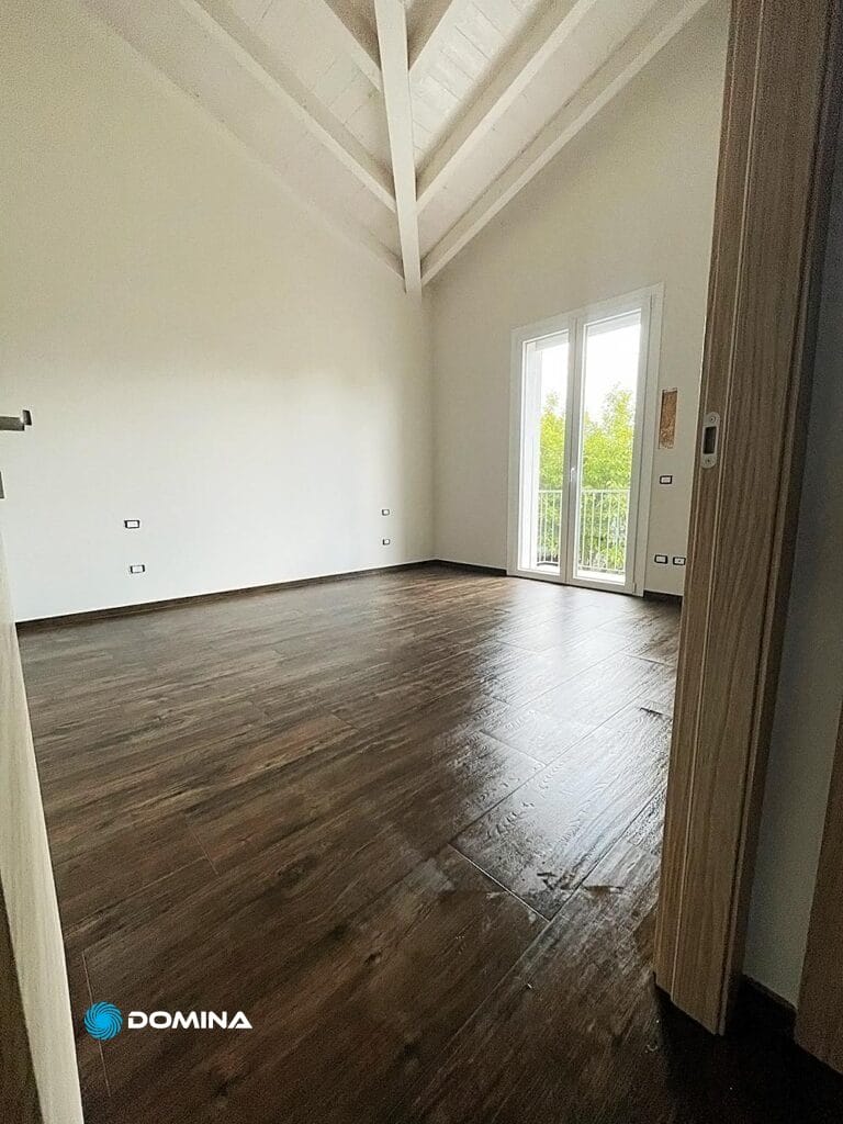 Una stanza vuota con pavimento in legno, pareti bianche e soffitto alto. Lo spazio incontaminato, risultante dalle belle pulizie del cantiere Malnate, ha una porta che si apre su un balcone con verde visibile all'esterno.