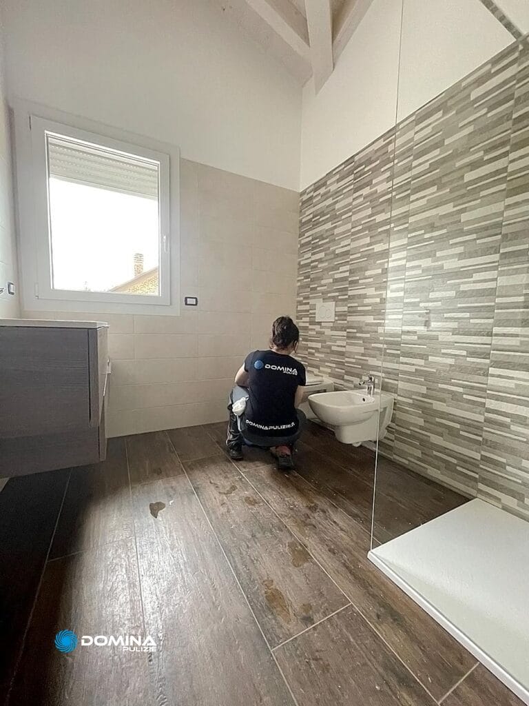 Una persona si accovaccia per lavorare all'installazione o alla manutenzione di un bidet sospeso in un bagno con pareti piastrellate e pavimento in legno, esemplificando l'attenzione ai dettagli vista in Domina Pulizie.
