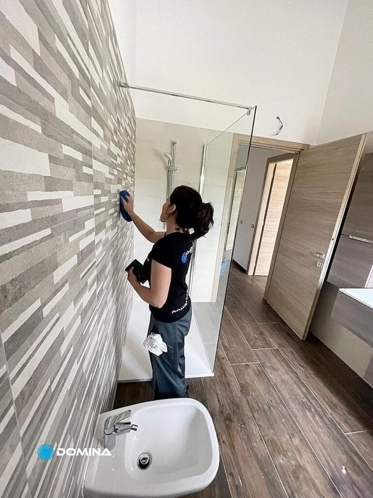 Una persona sta pulendo una parete piastrellata in un bagno moderno con cabina doccia in vetro, pavimento in legno e bidet. Con in mano un panno e una bomboletta spray, lavorano meticolosamente come se stessero eseguendo le pulizie del cantiere Malnate per Domina Pulizie.