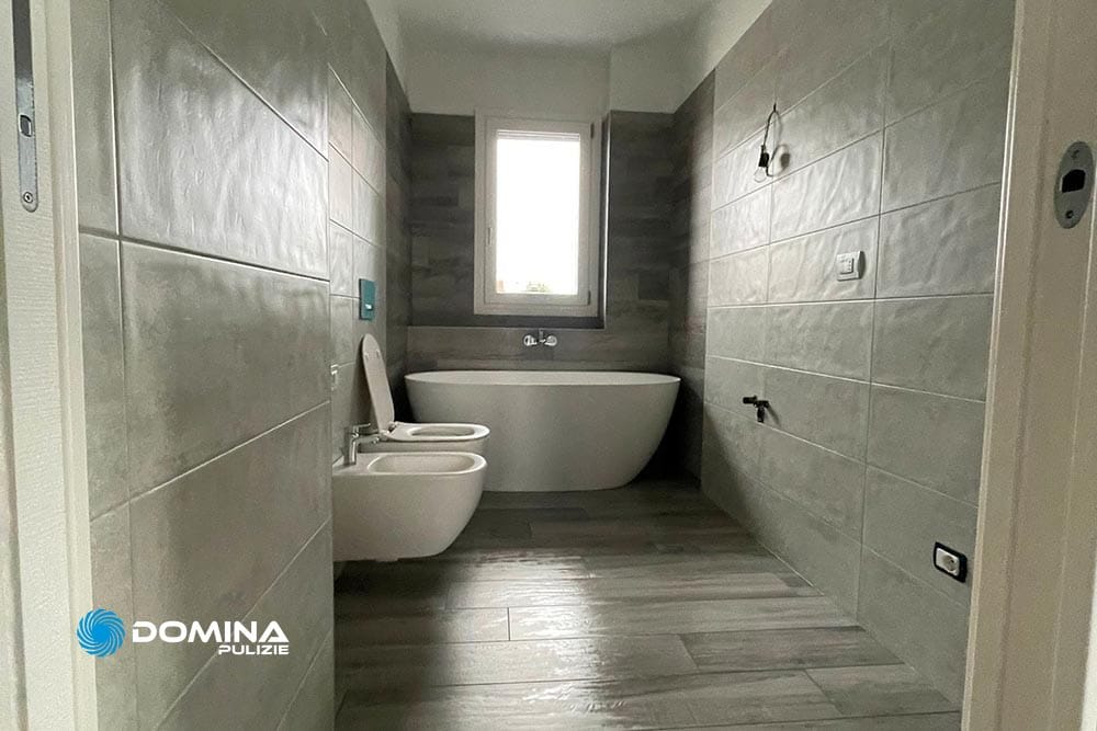 Bagno moderno con vasca freestanding, zona doccia separata, WC e bidet, con piastrelle grigie e finestra, curato meticolosamente da Domina Pulizie.