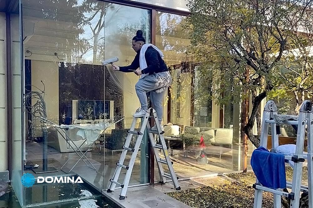 Persona in piedi su una scala che pulisce una grande finestra di una casa moderna, con alberi e un piccolo gioco d'acqua nelle vicinanze. All'interno, attraverso il vetro, tavolo e sedie riflettono le meticolose Pulizie Appartamenti di Servizi Professionali di Arcore.