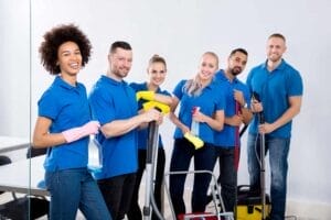 Un gruppo di sei individui sorridenti che indossano camicie blu sono riuniti a Milano, rappresentando un'impresa di pulizie, con in mano vari strumenti di pulizia come bottiglie spray, uno spazzolone e un aspirapolvere.