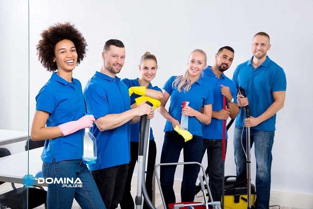 Un gruppo di sei individui sorridenti che indossano camicie blu sono riuniti a Milano, rappresentando un'impresa di pulizie, con in mano vari strumenti di pulizia come bottiglie spray, uno spazzolone e un aspirapolvere.