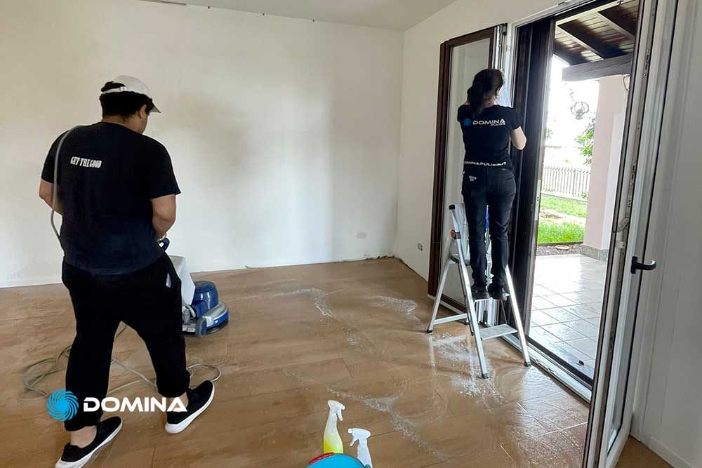 Due persone di Domina Pulizie stanno pulendo meticolosamente una stanza vuota a Malnate. Uno aziona una macchina lavapavimenti sul pavimento in legno, mentre l'altro sta su una scala, facendo scintillare la porta di vetro. La porta aperta sul patio aggiunge un tocco di luminosità agli sforzi post-ristrutturazione.