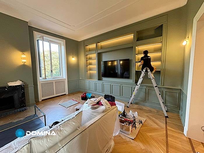 Una persona in piedi su una scala regola i ripiani di un mobile da incasso in un soggiorno. Nelle vicinanze sono posizionati un divano coperto da un telo di plastica e vari attrezzi. La camera, parte di un appartamento di prestigio nel centro storico di Milano, ha pareti verdi e pavimento in legno.