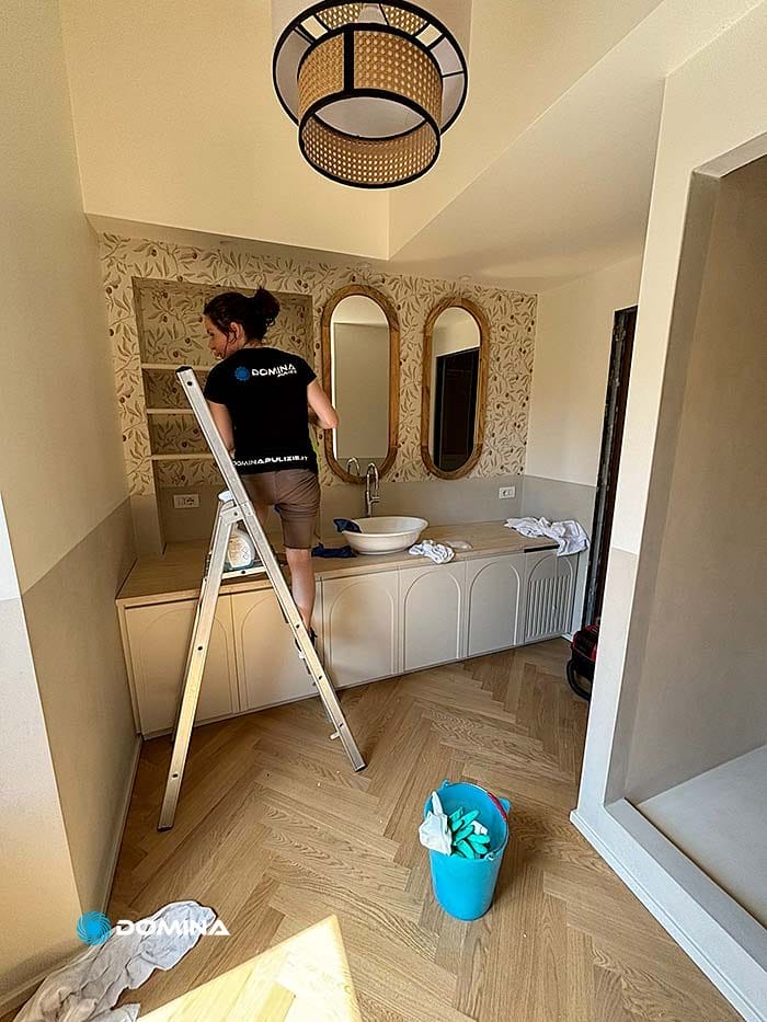 Una persona in piedi su una scala pulisce la parete del bagno vicino a un lavandino, con un secchio blu di prodotti per la pulizia sul pavimento. Il lussuoso bagno di questo appartamento nel centro storico di Milano è dotato di due specchi e carta da parati decorativa.