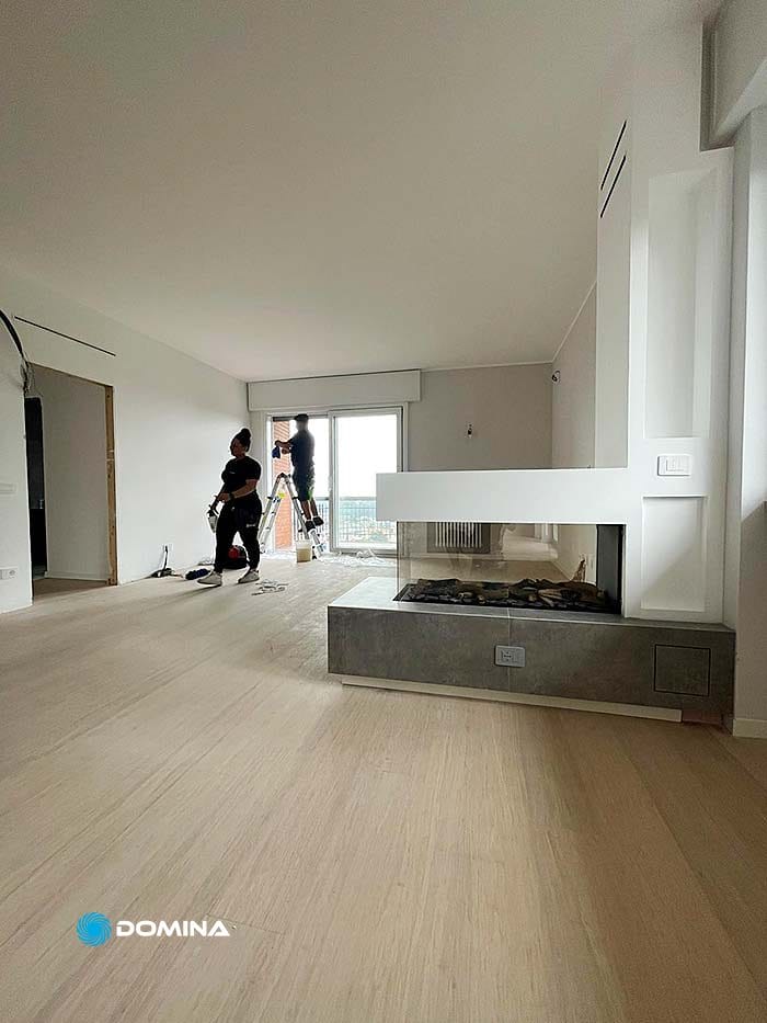 Due persone lavorano alla ristrutturazione della casa in una stanza luminosa, parzialmente rifinita, con pavimento in legno chiaro e caminetto in muratura, pronta per le pulizie post cantiere Busto Arsizio per farla risplendere.