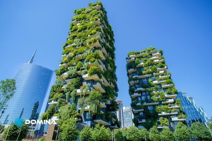 Due moderni grattacieli ricoperti da una vegetazione lussureggiante si stagliano contro il cielo azzurro di Milano, con grattacieli circostanti e alberi sullo sfondo.