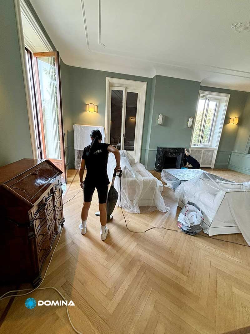 Una persona sta aspirando un pavimento di legno in una stanza con mobili coperti da lenzuola. Le pareti azzurre e le due grandi finestre donano allo spazio un'atmosfera ariosa. Sembra che la migliore Impresa di Pulizie di Milano, nota per i suoi Servizi Professionali di prim'ordine, sia stata al lavoro qui.